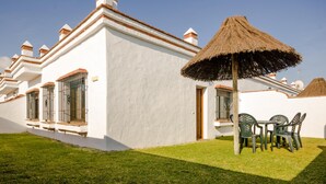 A white building with a thatched roof, a small patio with green plastic chairs, and a well-maintained lawn.