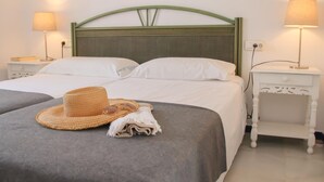 Ein ordentlich bezogenes Bett mit weißen Leinentüchern, ein grüner Kopfteil und ein gewebter Hut auf dem Bett. Zwei Nachttische mit Lampen stehen bereit, darüber hängt ein gerahmtes Kunstwerk.