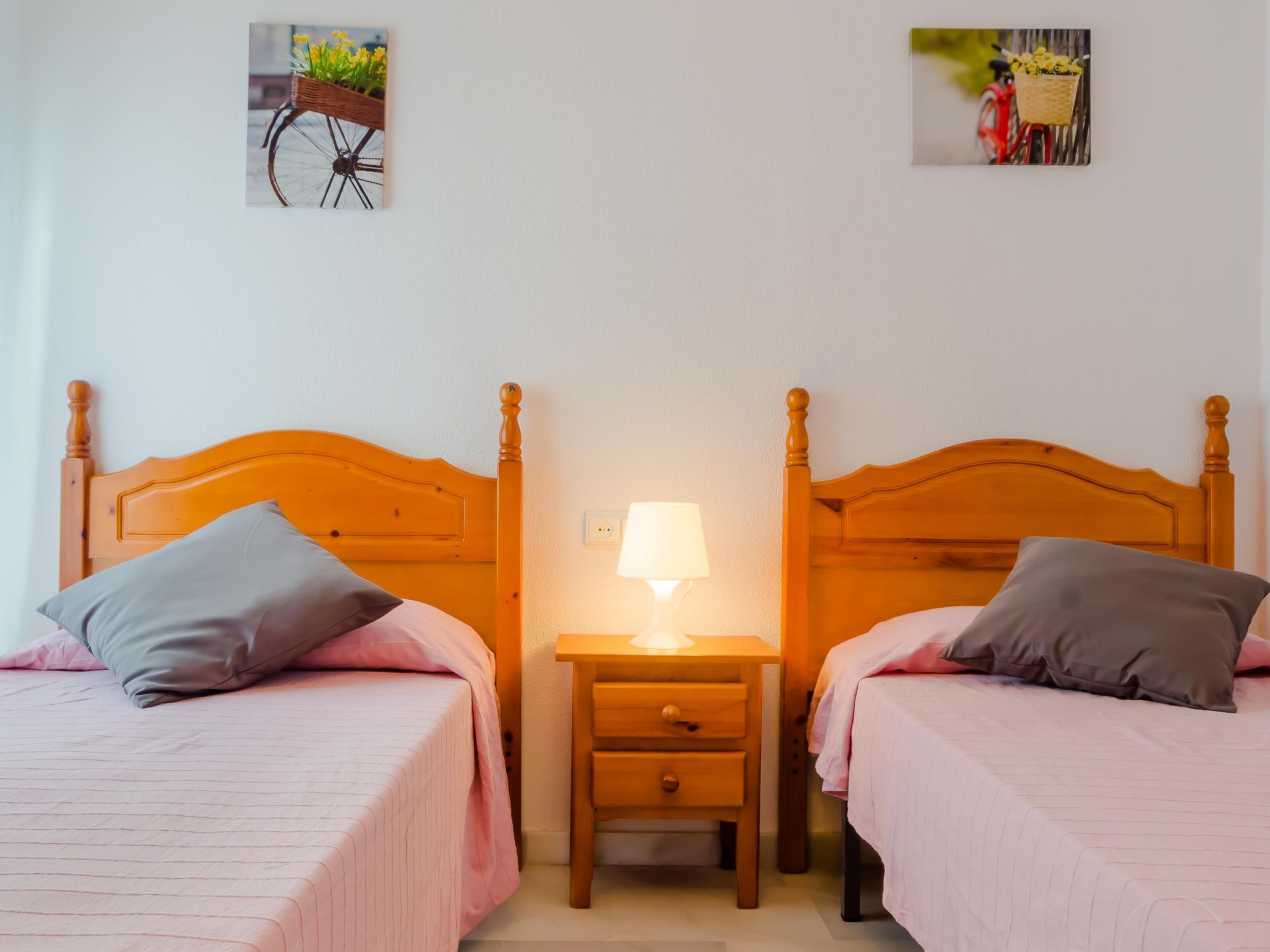 Habitación doble con dos camas, cada una con cabecera de madera, mesita de noche con lámpara y un cuadro colgado en la pared de una bicicleta.