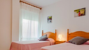 Ein Zimmer mit zwei Betten, jeweils mit einem Holzkopfstück, einem Nachttisch mit Lampe und einem Fenster mit durchsichtigen Vorhängen.