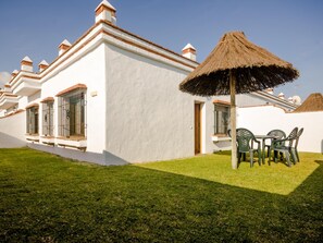 Un bâtiment blanc avec un toit de chaume, un petit patio avec des chaises en plastique vertes et une pelouse bien entretenue.