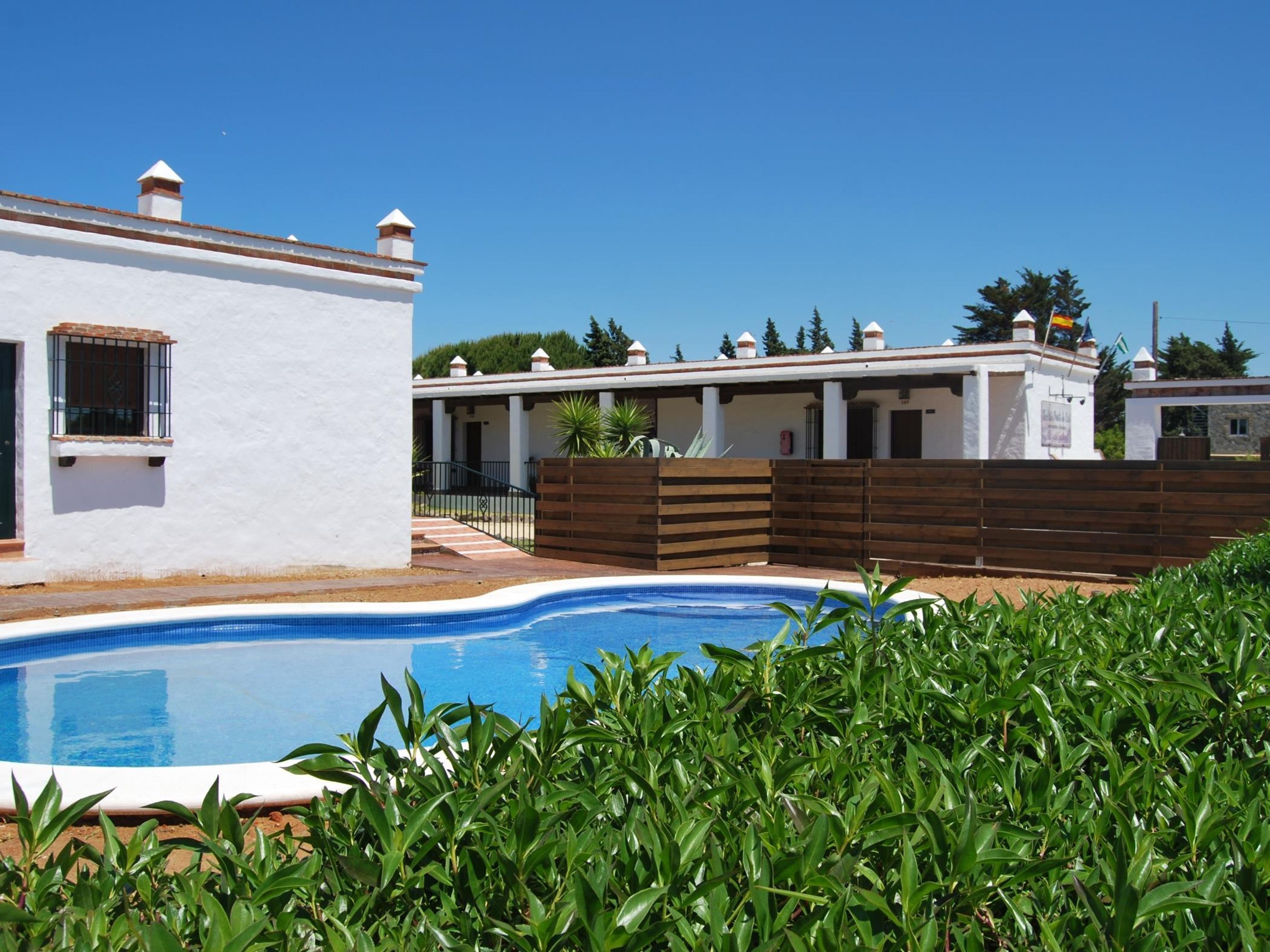 A pool area with a white building, wooden fence, and greenery.