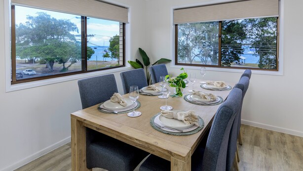 Comedor con una mesa de madera dispuesta para cuatro, ventanas amplias con persianas y vistas al exterior.