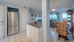 A kitchen with a stainless steel refrigerator, a granite countertop, and a dining area with wooden chairs. - Beachfront Condo w/ Ocean View, 2 Pools, Boardwalk to the Beach | Island Getaway (Port Aransas)
