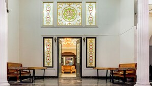 A lobby with a chandelier, two wooden benches, and three stained glass windows with intricate designs.