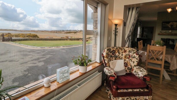 A cozy room with a large window offering a view of a rural landscape. A floral armchair is positioned near the window, and a small table with a lamp and a framed picture is visible.