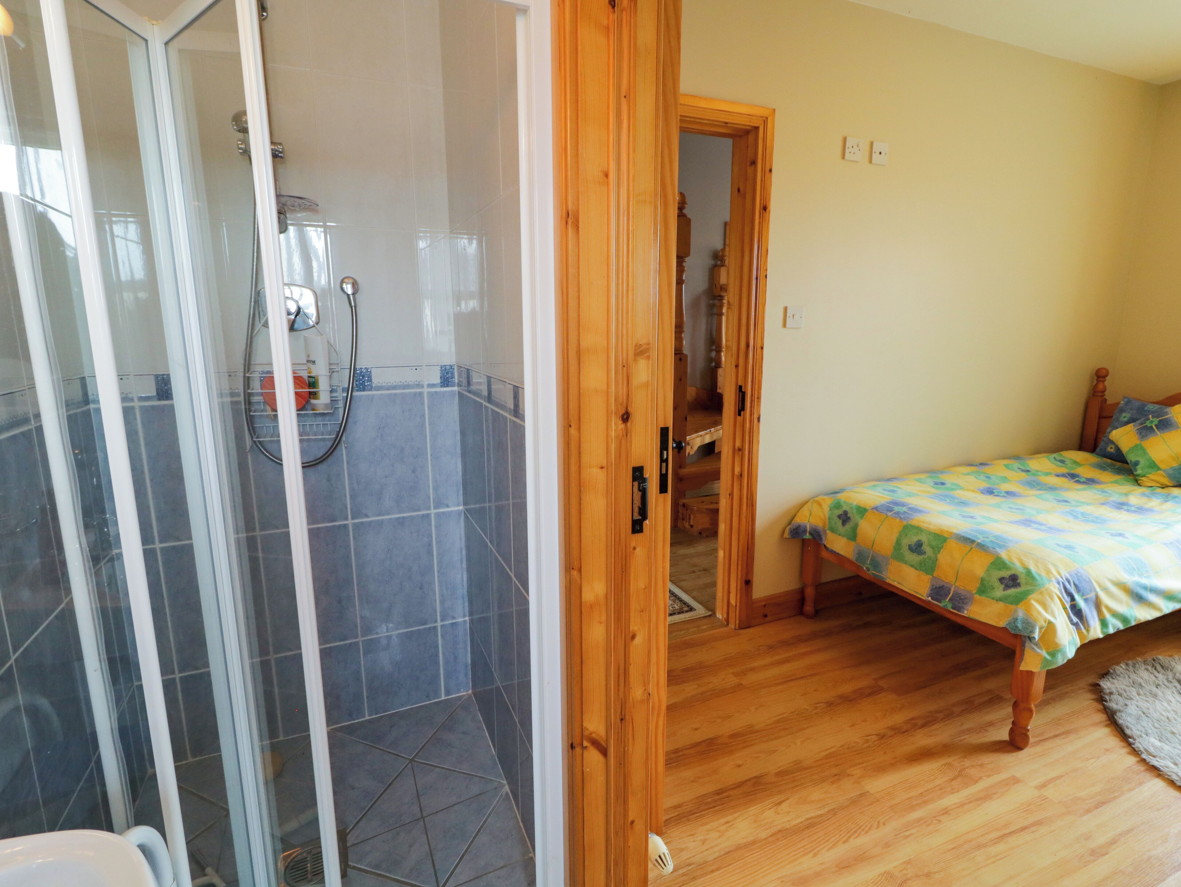 A room with a shower, wooden flooring, a bed with a colorful bedspread, and a wooden door.