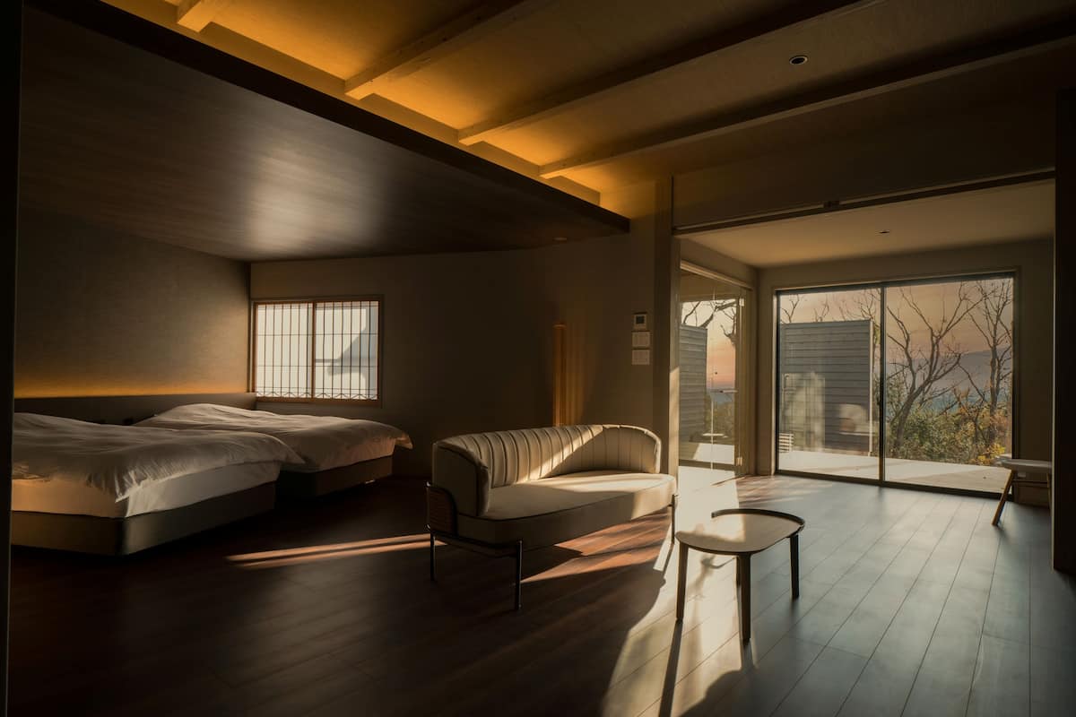 A modern bedroom with a large bed, a sofa, and a small table. There is a window with bars and a view of the outside.