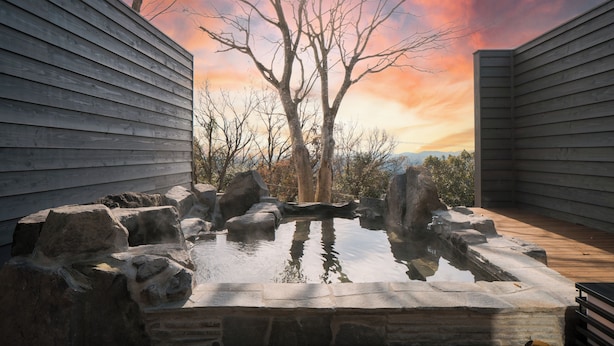 A hot tub surrounded by rocks and trees, with a wooden deck and a sunset in the background. - Kirinosato (Kirishima)