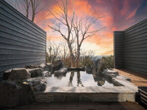 A hot tub surrounded by rocks and trees, with a wooden deck and a sunset in the background. - Kirinosato (Kirishima)