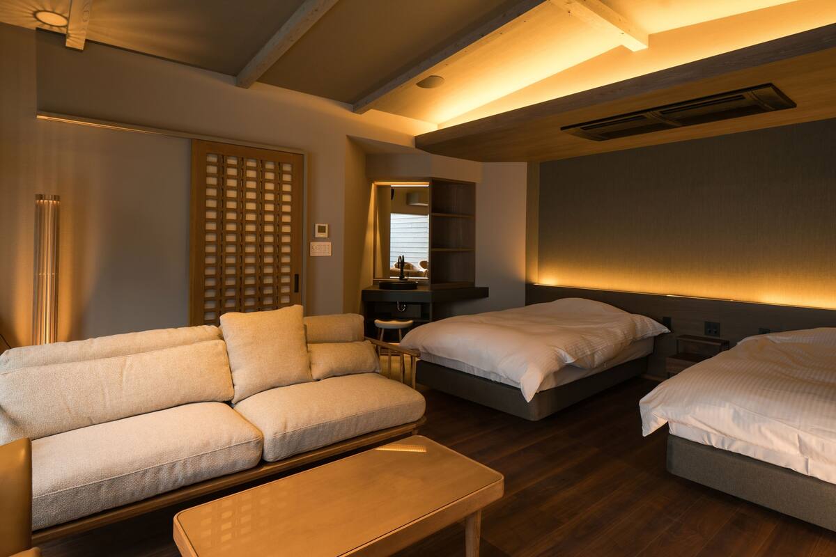 A modern hotel room with a sofa, two beds, a small table, and a wooden ceiling.