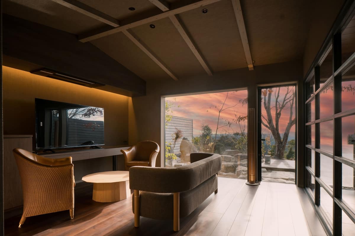 A modern living room with a flat-screen TV, wicker chairs, a round coffee table, and a large window offering a view of the outdoors.
