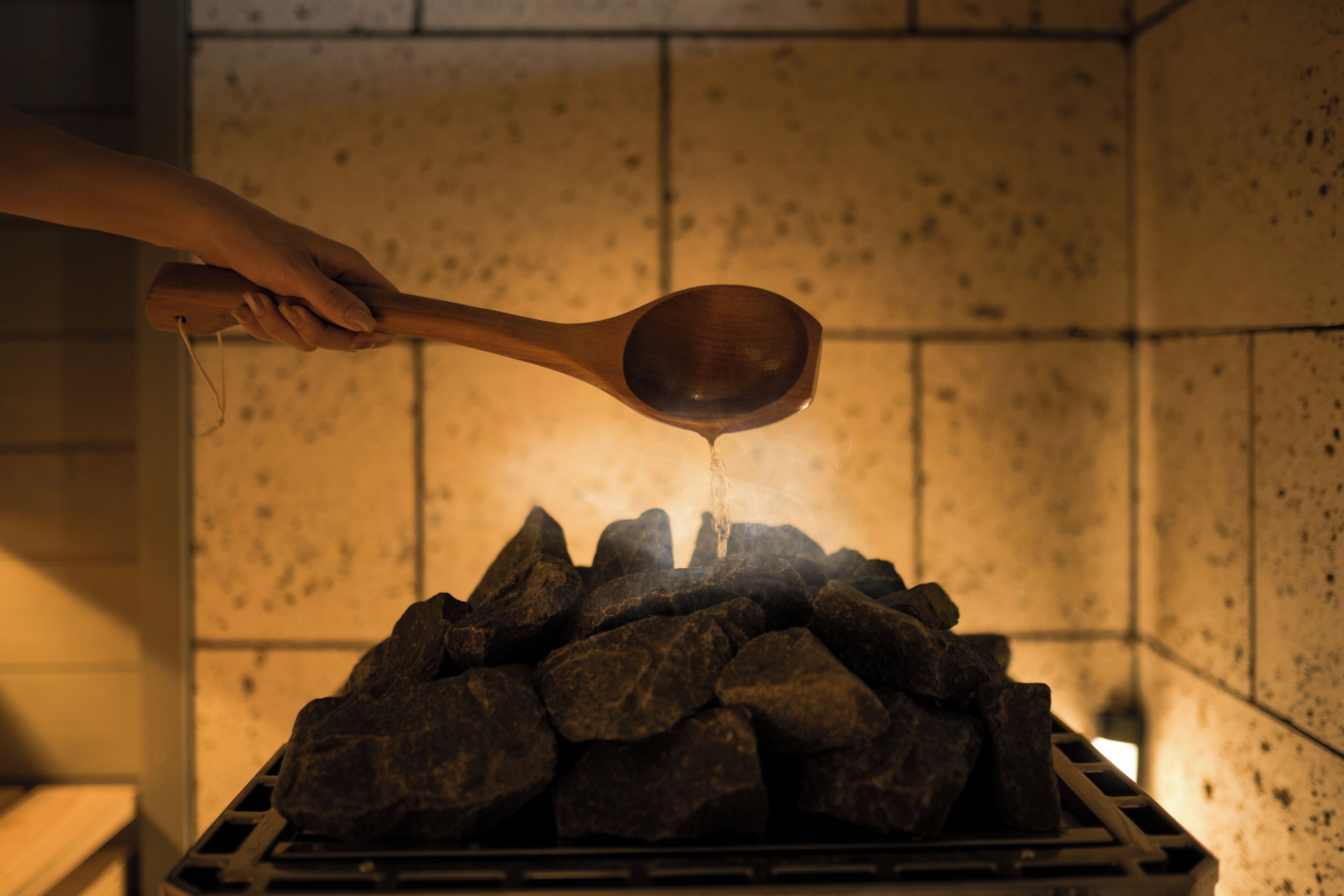 A hand holding a wooden ladle over a grill with charcoal.