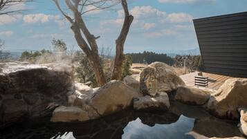 Un área de aguas termales con rocas grandes, una pasarela de madera y vistas a árboles y montañas al fondo.