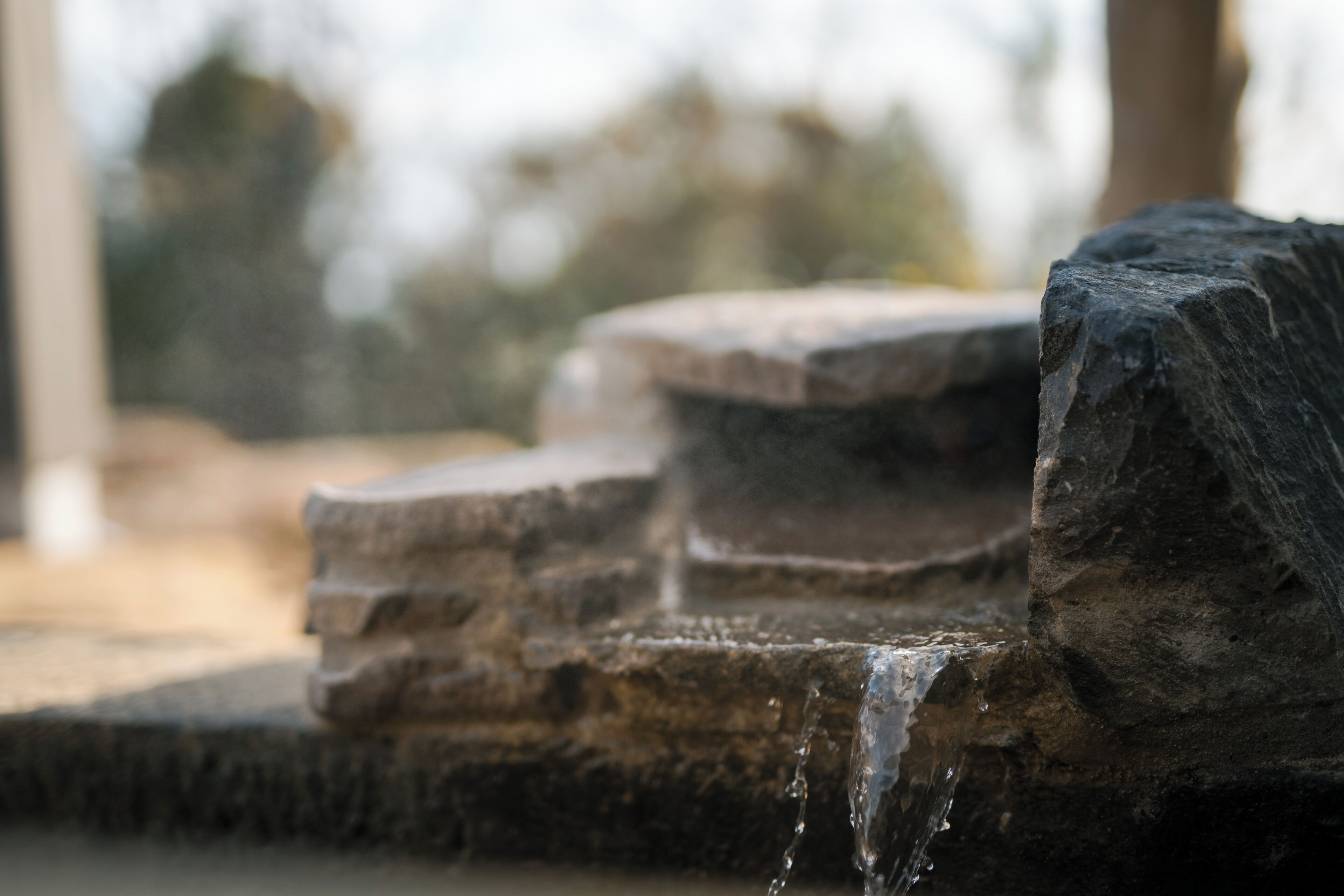 Una fuente de piedra con agua en cascada, sobre un fondo natural desenfocado.