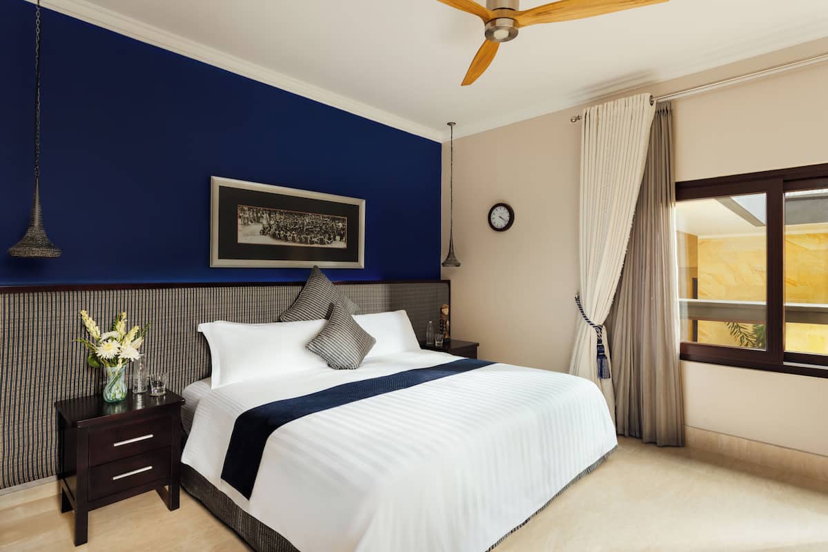 A hotel room with a large bed, a nightstand with a vase of flowers, a framed picture on the wall, a ceiling fan, a clock, and a window with curtains.