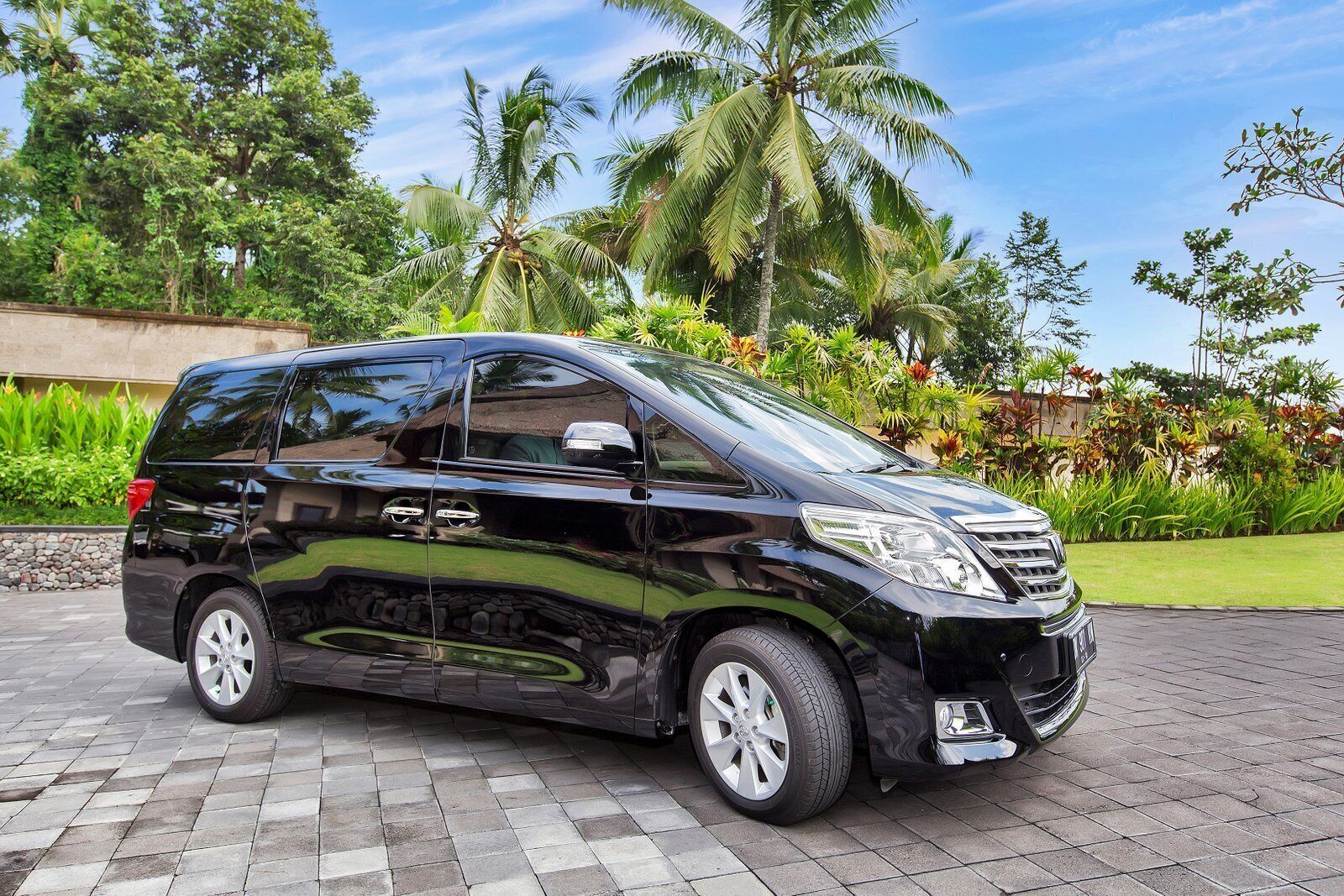 A black minivan parked on a paved area with a garden and palm trees in the background.
