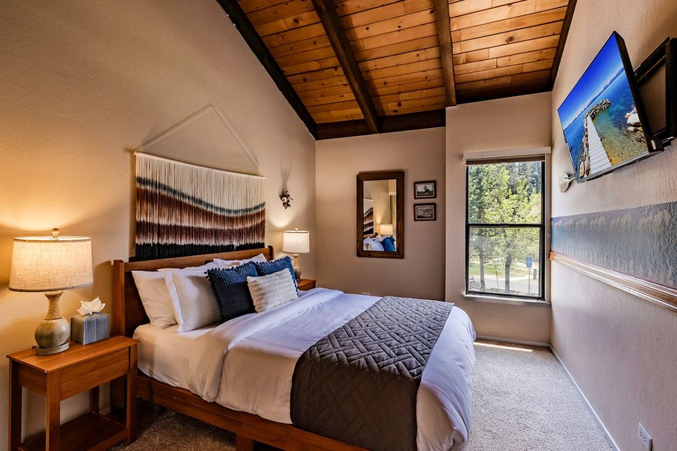 Une chambre avec un plafond en bois, un lit recouvert de plusieurs oreillers, une table de chevet avec une lampe, un miroir fixé au mur et une fenêtre donnant sur la forêt.