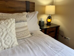 Ein ordentlich bezogenes Bett mit weißen Leinentextilien und dekorativen Kissen, ein Nachttisch mit Lampe sowie ein Holz-Kopfteil.
