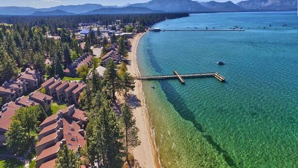 An aerial view of a coastal area with a pier extending into a clear blue lake, surrounded by residential buildings and dense forest.