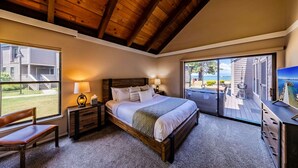 A bedroom with a wooden ceiling, a large bed, a nightstand with a lamp, a chair, and a view of the outside.
