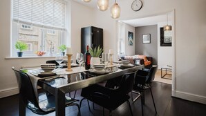 A dining area with a long table set for four, black chairs, hanging pendant lights, and a view of the street through large windows. - Modern contemporary dog friendly - Licata's Apartment (Cirencester)
