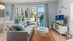 Una sala de estar moderna con un sofá gris, una mesa de centro blanca y un televisor sobre un mueble blanco. Se aprecia un amplio ventanal con vistas a un edificio y zonas verdes.