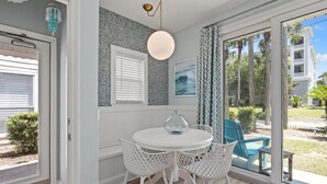Un comedor con una mesa y sillas blancas, una lámpara colgante y vistas a un edificio y follaje exterior.