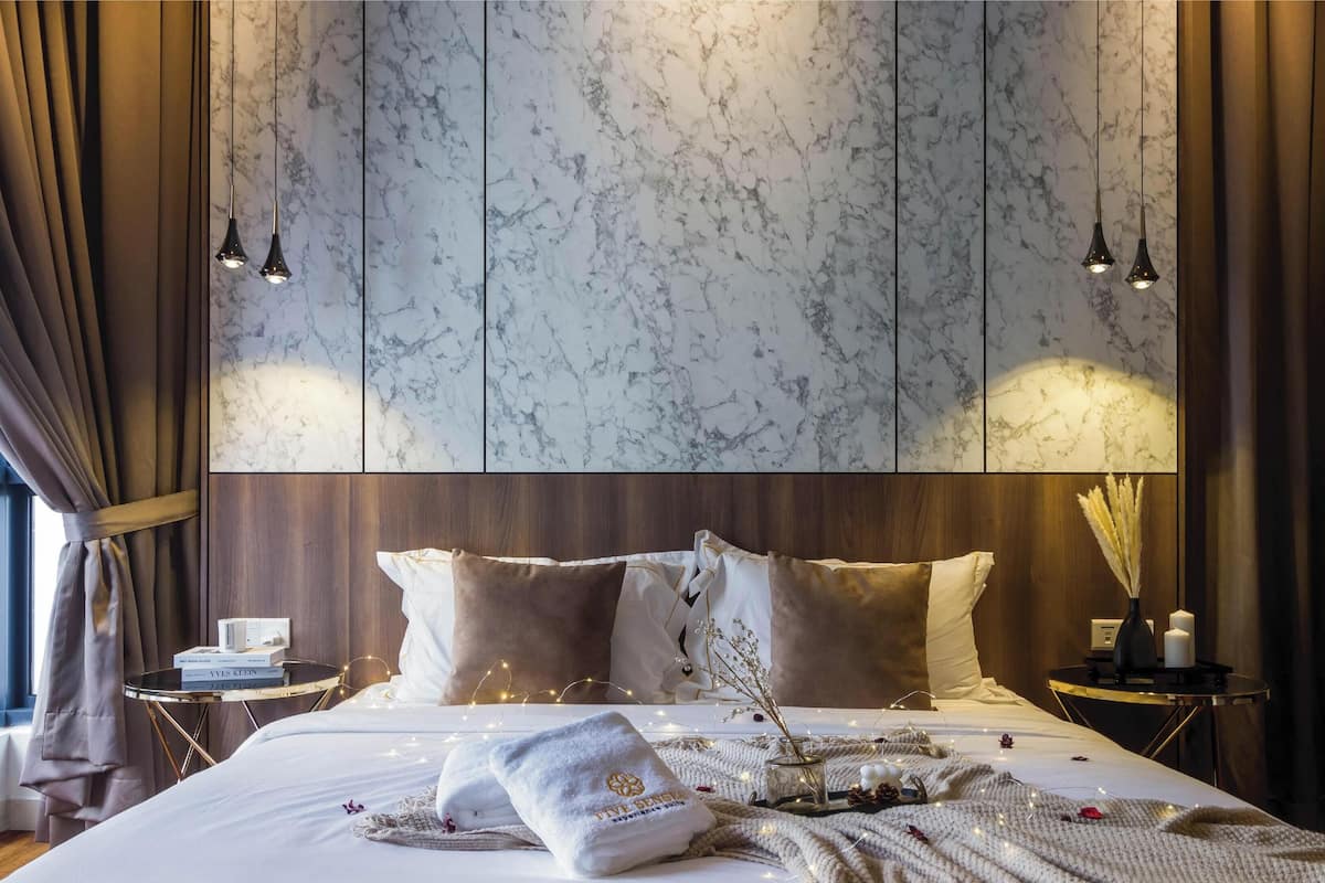 Un dormitorio moderno con un cabecero estampado con diseño de mármol, una cama bien tendida con una colcha blanca y una mesita de noche con un libro y una lámpara.