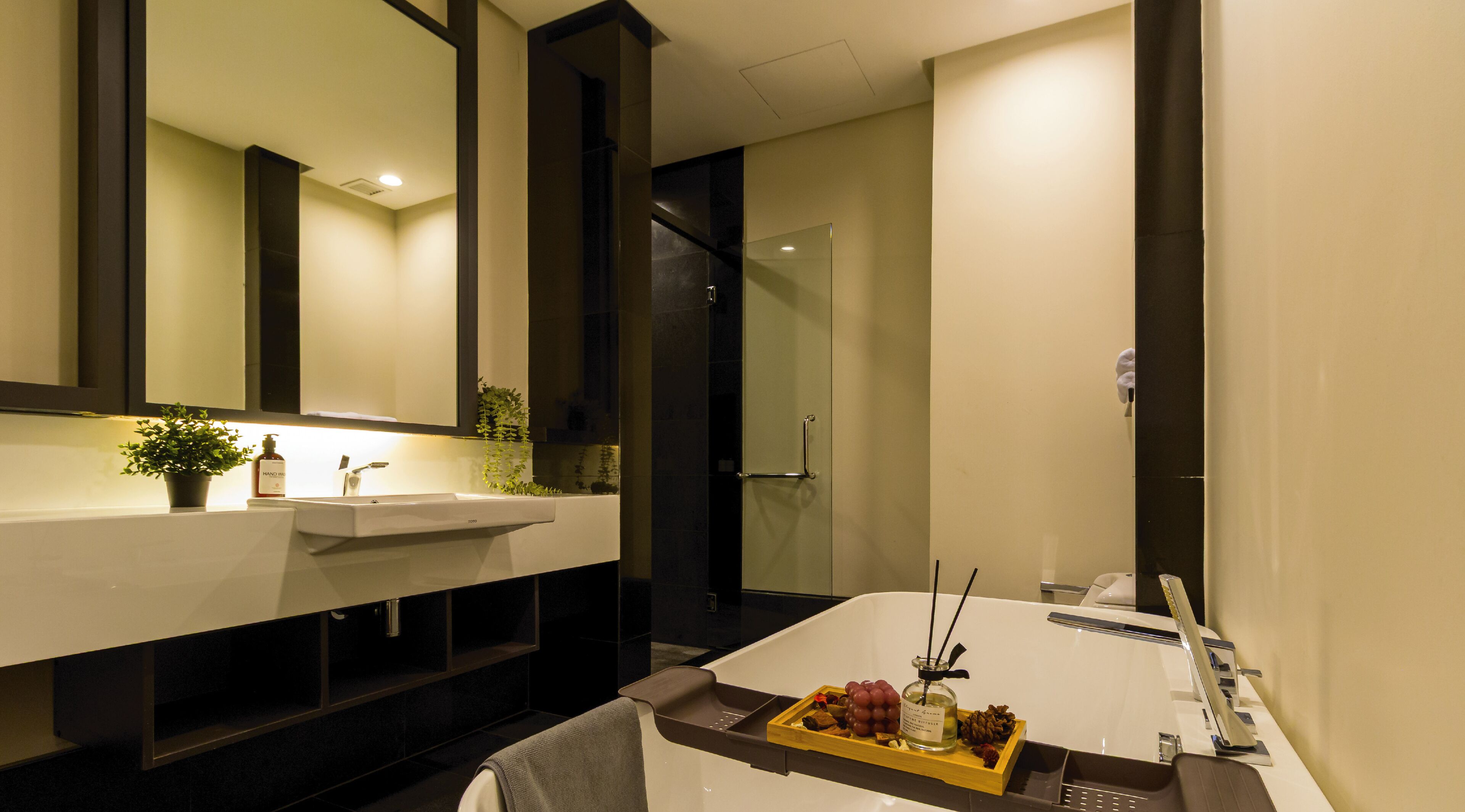 A modern bathroom with a large bathtub, a sink, and a mirror. There is a tray with toiletries and a bottle of hand soap on the sink.