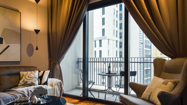 Un salon moderne avec un canapé, un fauteuil et une petite table sur laquelle se trouvent une bouteille et des objets décoratifs. Une grande fenêtre, ornée de rideaux, offre une vue sur un immeuble doté de nombreuses fenêtres et de balcons.