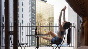 Une personne est assise sur une chaise de balcon, devant une table, avec une bouteille de vin, et elle a devant elle une vue sur un paysage urbain avec de hauts immeubles.