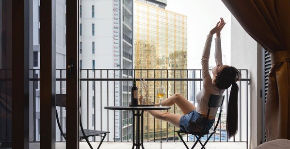 A person sitting on a balcony chair with a table and a bottle of wine, overlooking a cityscape with tall buildings. - The Mews KLCC by Five Senses (Kuala Lumpur)