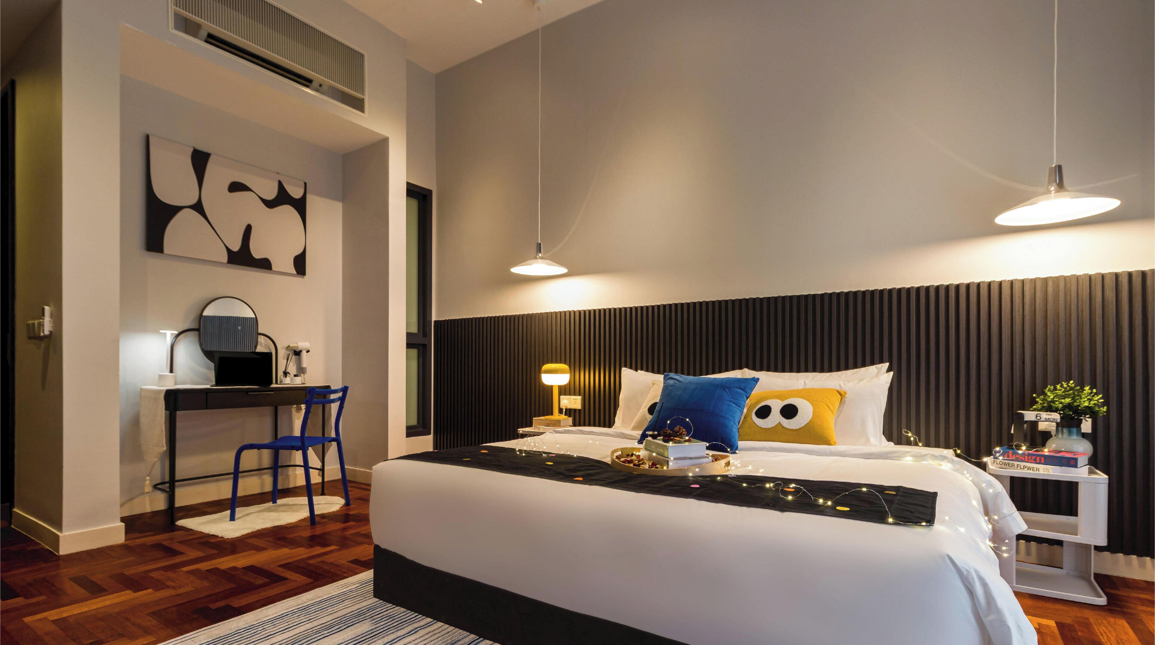 A modern hotel room with a large bed, bedside table, lamp, and a desk with a chair.