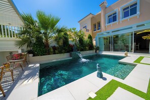 A swimming pool with a waterfall feature, surrounded by lush greenery and a patio with outdoor furniture. - Luxury Short Term Stay 2BHK plus Maids - RANCHES (Dubai)