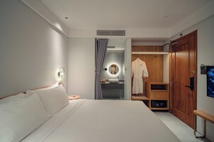 A modern hotel room with a large bed, a vanity with a mirror, a wooden wardrobe, and a bench.