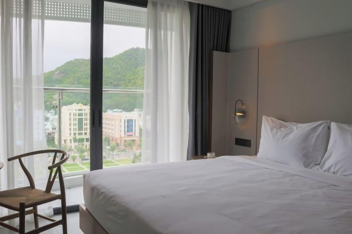 A hotel room with a large bed, a chair, and a window offering a view of buildings and greenery.