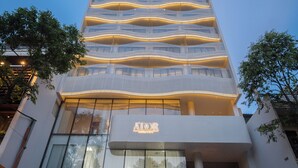 Un hôtel moderne, reconnaissable à son grand panneau « AIOOR », doté de nombreux balcons et devant lequel est garé un SUV jaune.