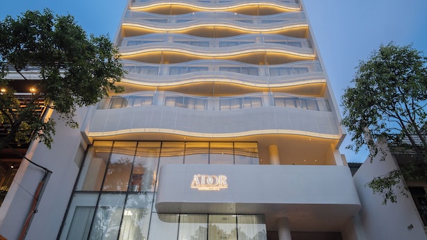 Un hôtel moderne, reconnaissable à son grand panneau « AIOOR », doté de nombreux balcons et devant lequel est garé un SUV jaune.