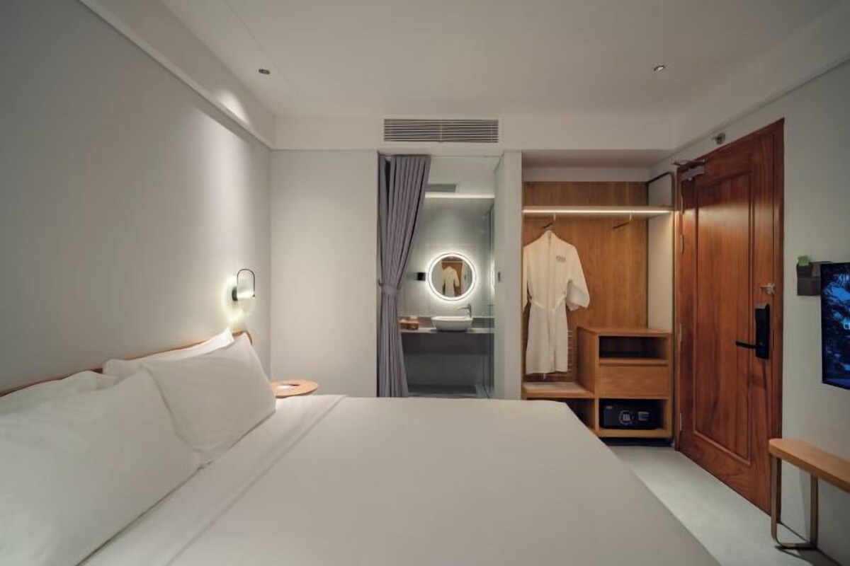 A modern hotel room with a large bed, a walk-in closet, a vanity with a mirror, and a bench by the door.