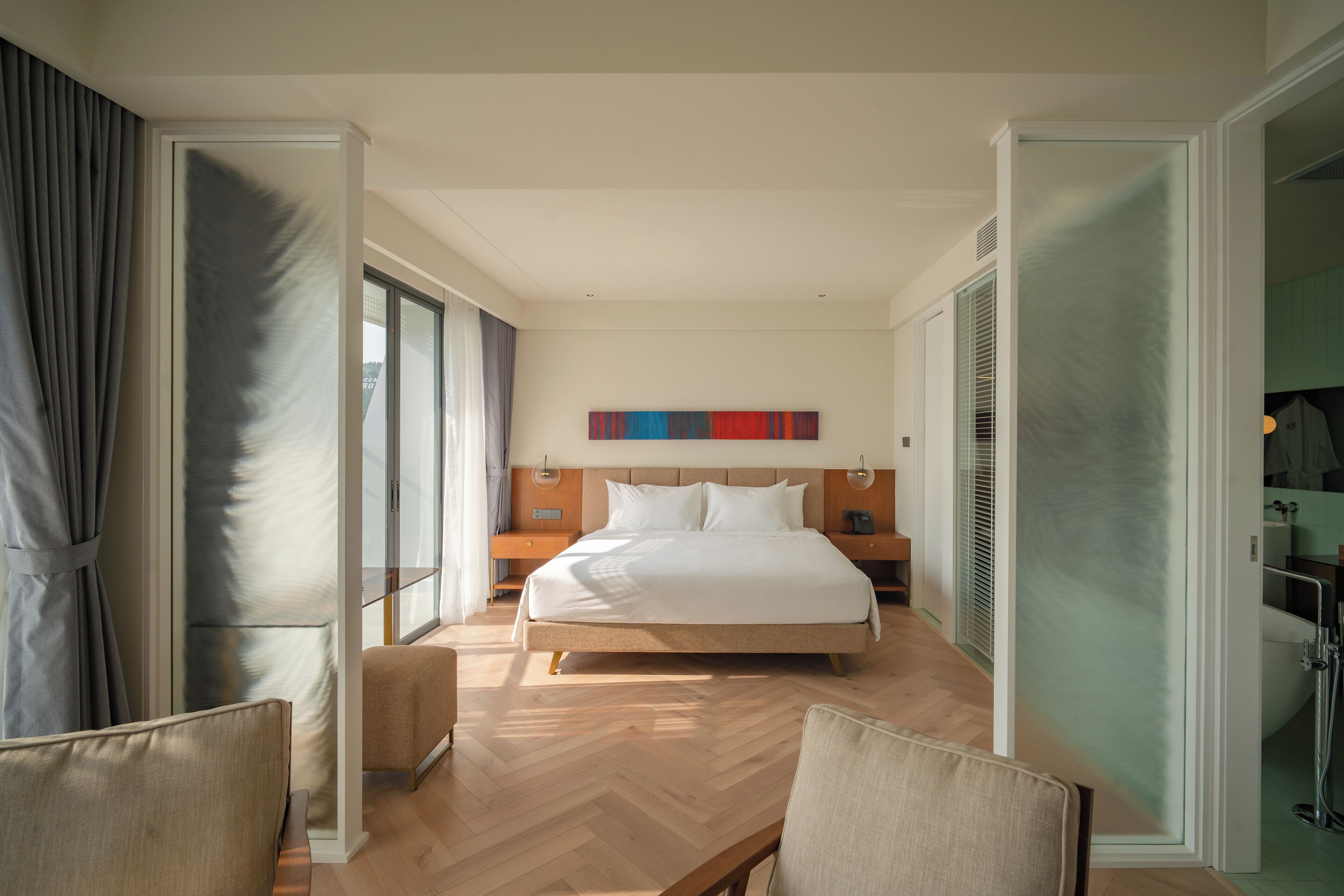 A hotel room with a large bed, a bedside table, a chair, and a view of the city through the window.