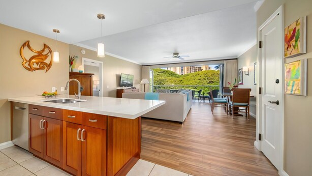A modern kitchen with wooden cabinets and a large island, a dining area with a table and chairs, and a living room with a sofa and a TV. - Honua Kai Konea 342 Partial Ocean View 2 BD 2BA (Lahaina)