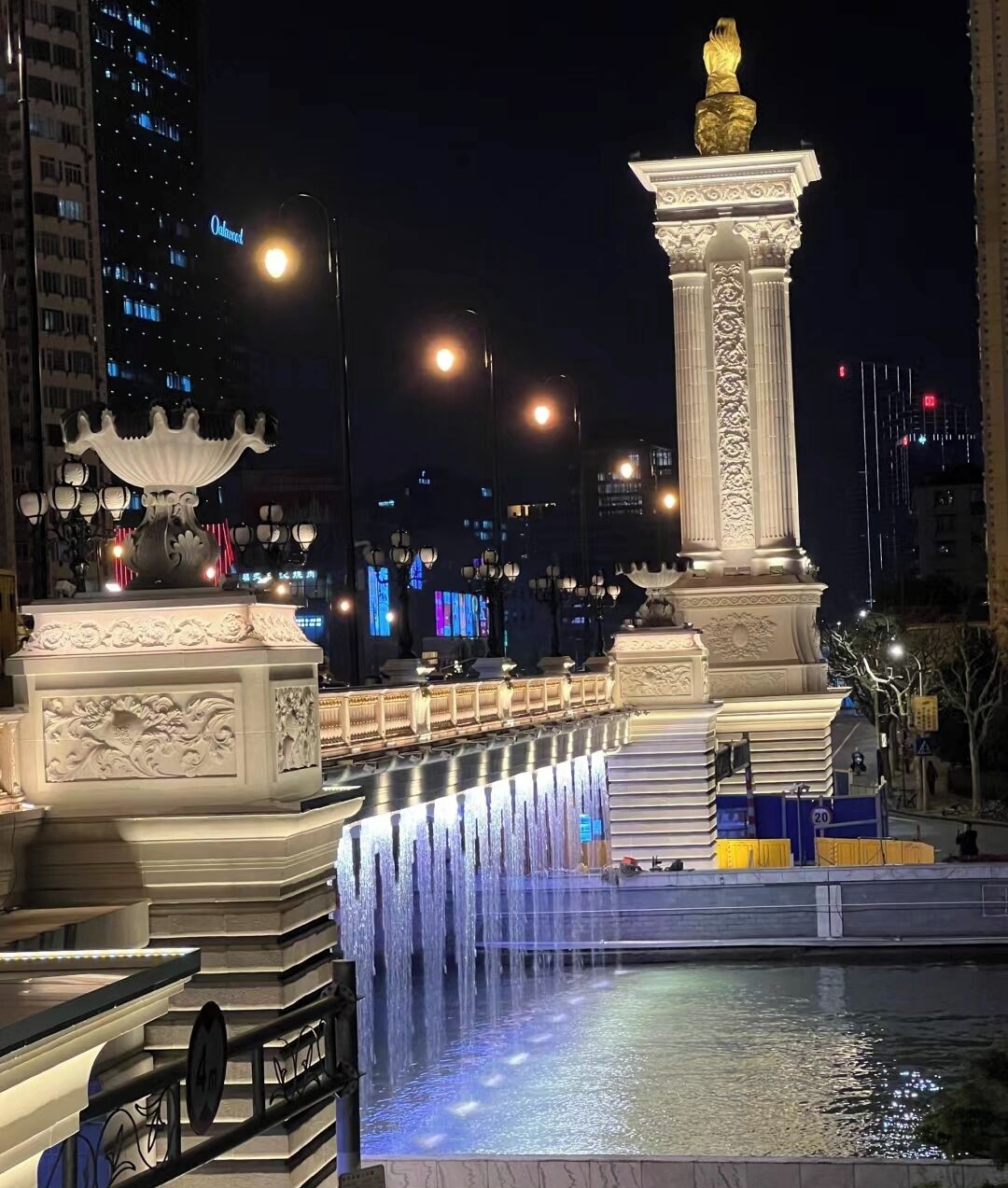 Un paisaje urbano nocturno con edificios iluminados, una fuente y una estatua.