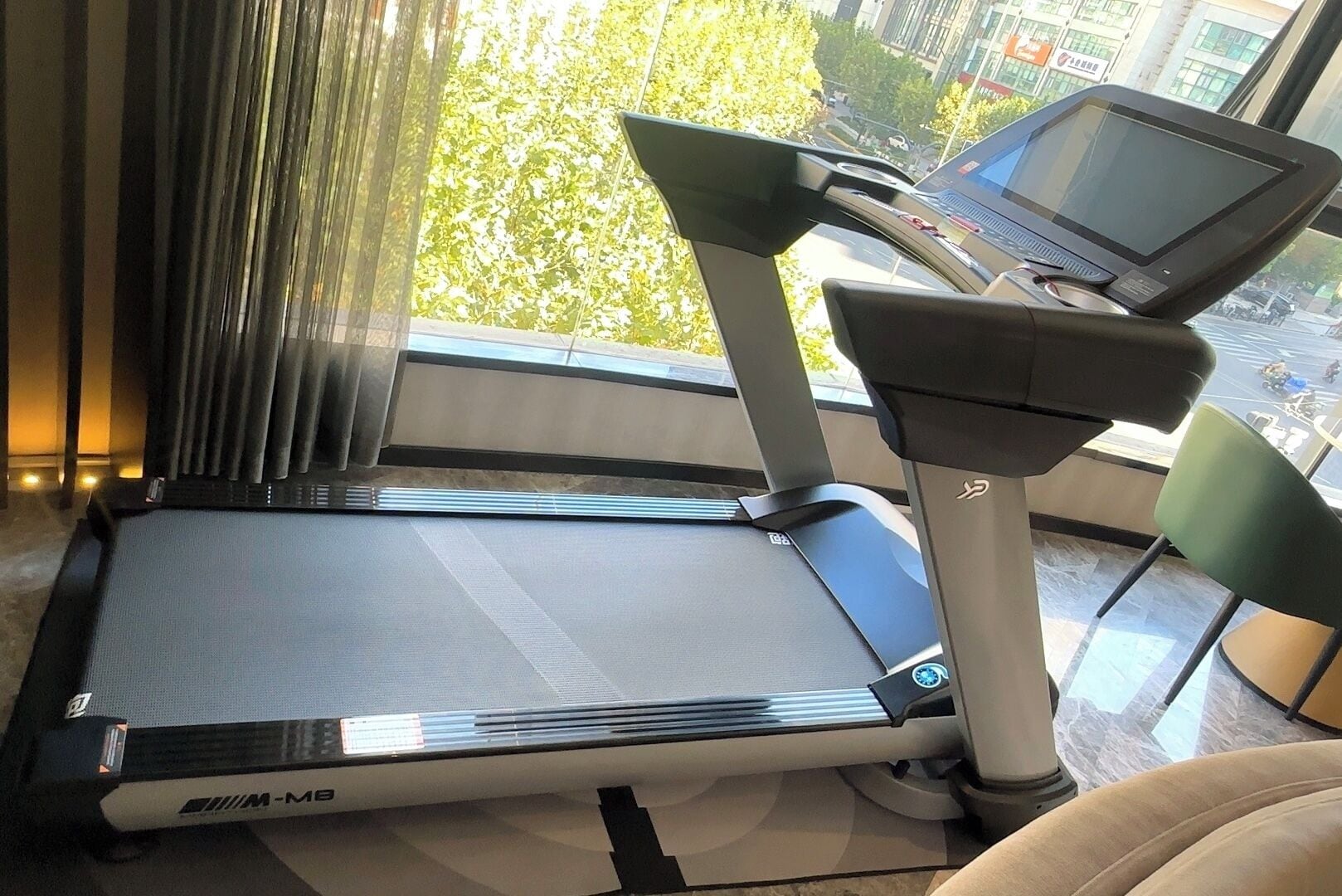 A treadmill with a digital display, positioned near a window with curtains, and a chair in the foreground.