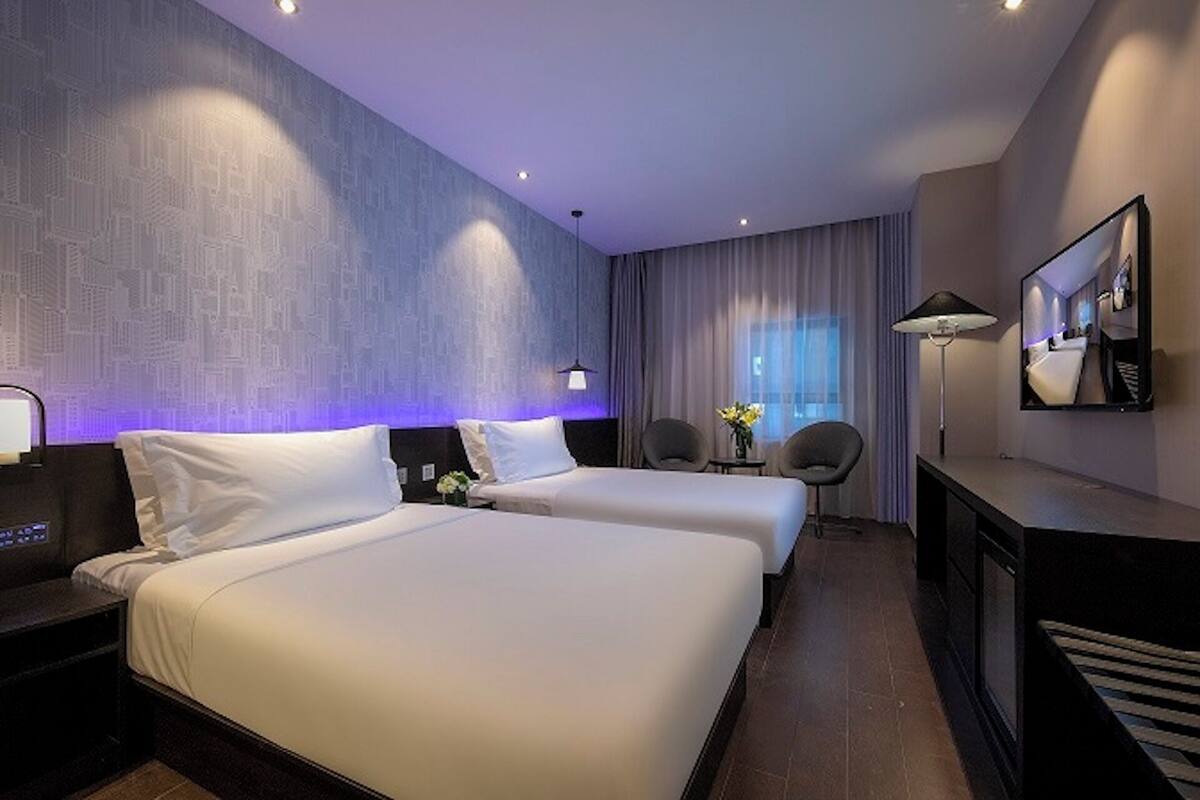 A hotel room with two beds, a desk, a chair, a lamp, and a large window with curtains.