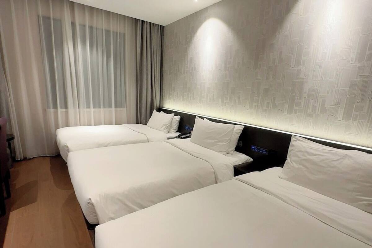 A hotel room with two beds, a wooden floor, a large window with curtains, and a wall with a subtle pattern.