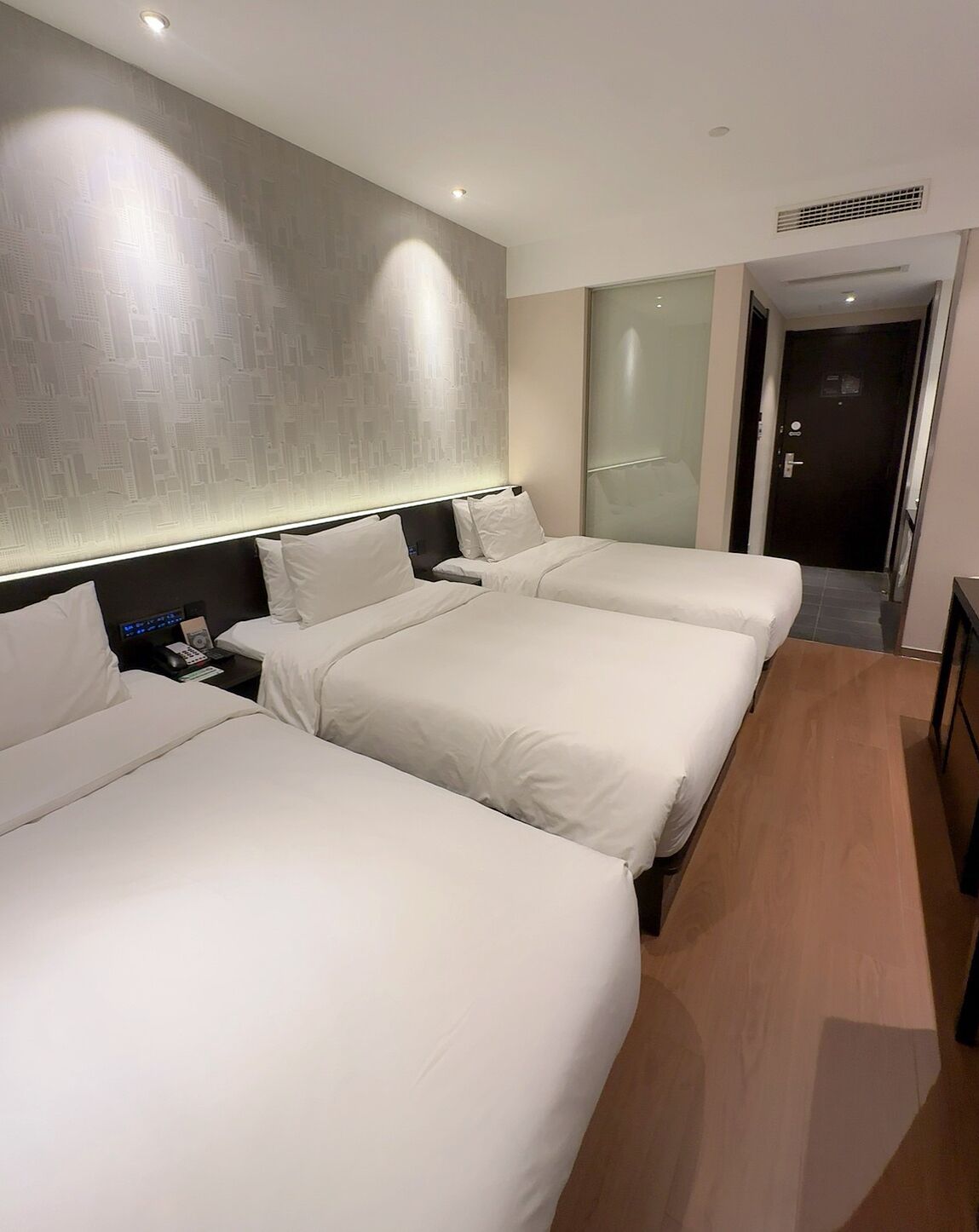 Habitación de hotel con dos camas, una mesita de noche, una lámpara de pared y una puerta que da a otra habitación.