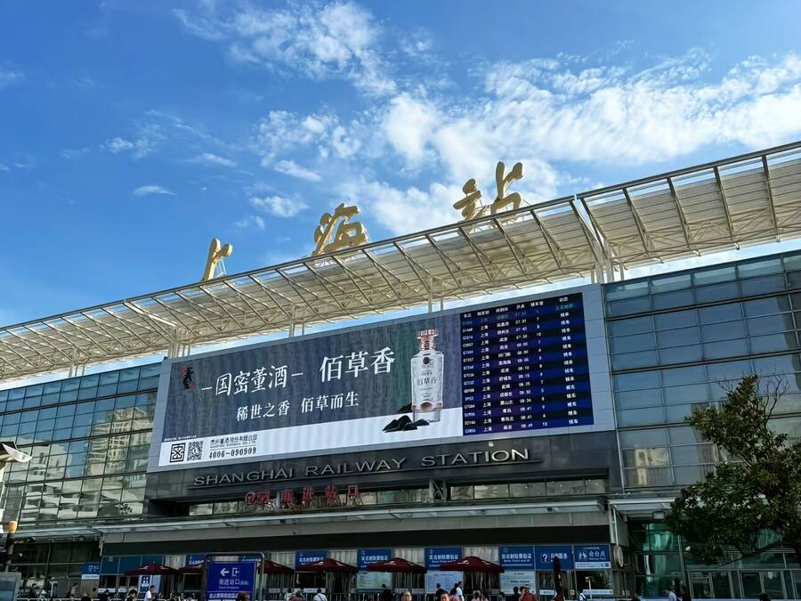A train station with a large digital display, glass facade, and a clear sky.