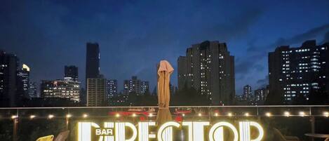 A rooftop bar with a curved sofa and a round table, overlooking a city skyline at night.