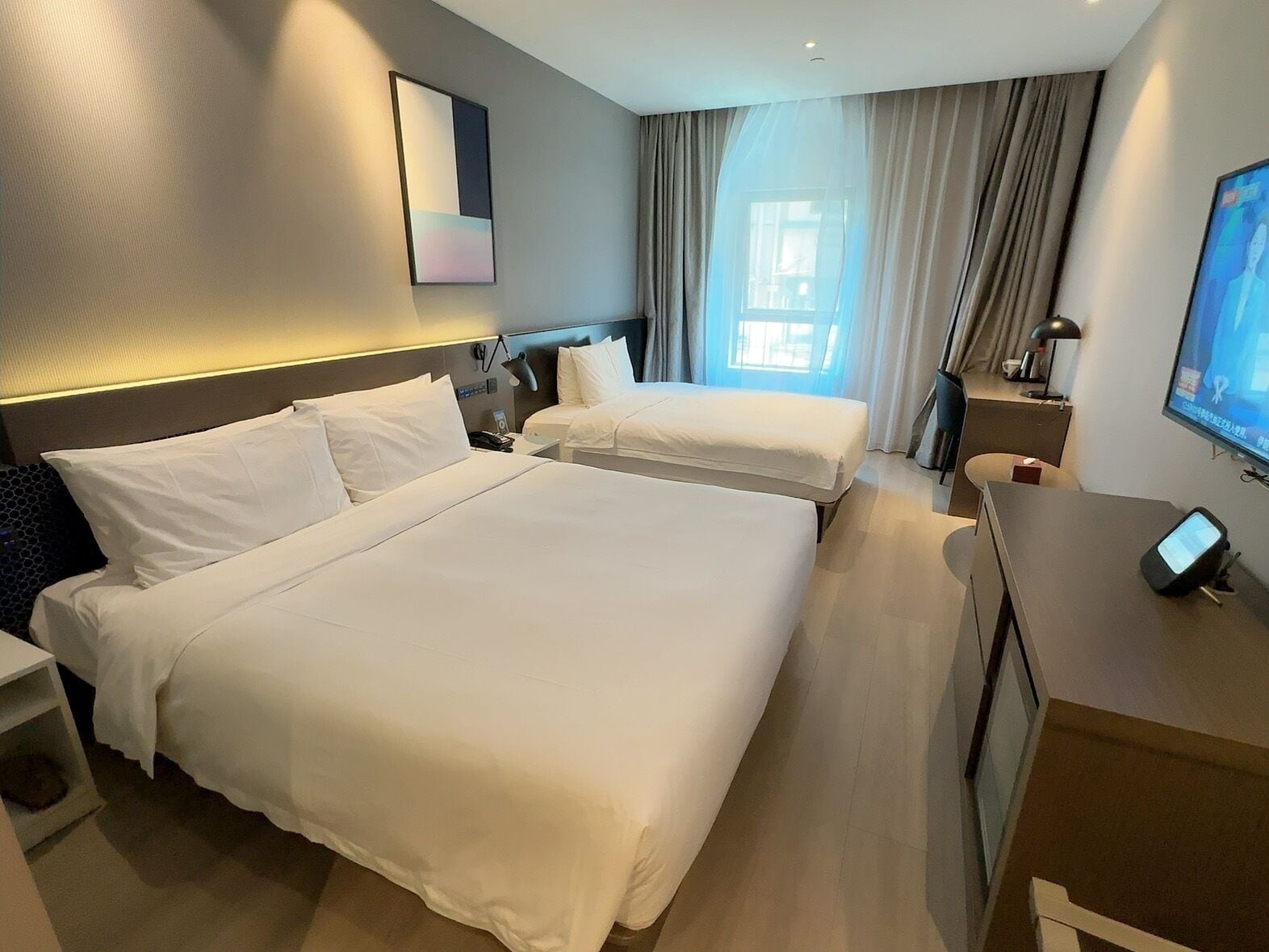 A hotel room with two beds, a desk, a TV, and a window with curtains.